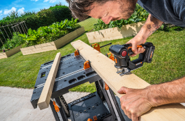 Man using a jigsaw on wooden plank in garden; DIY woodworking, outdoor carpentry, home improvement.
