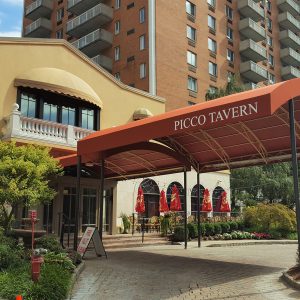 Picco Tavern entrance with red canopy, outdoor seating, lush greenery, and residential building backdrop.