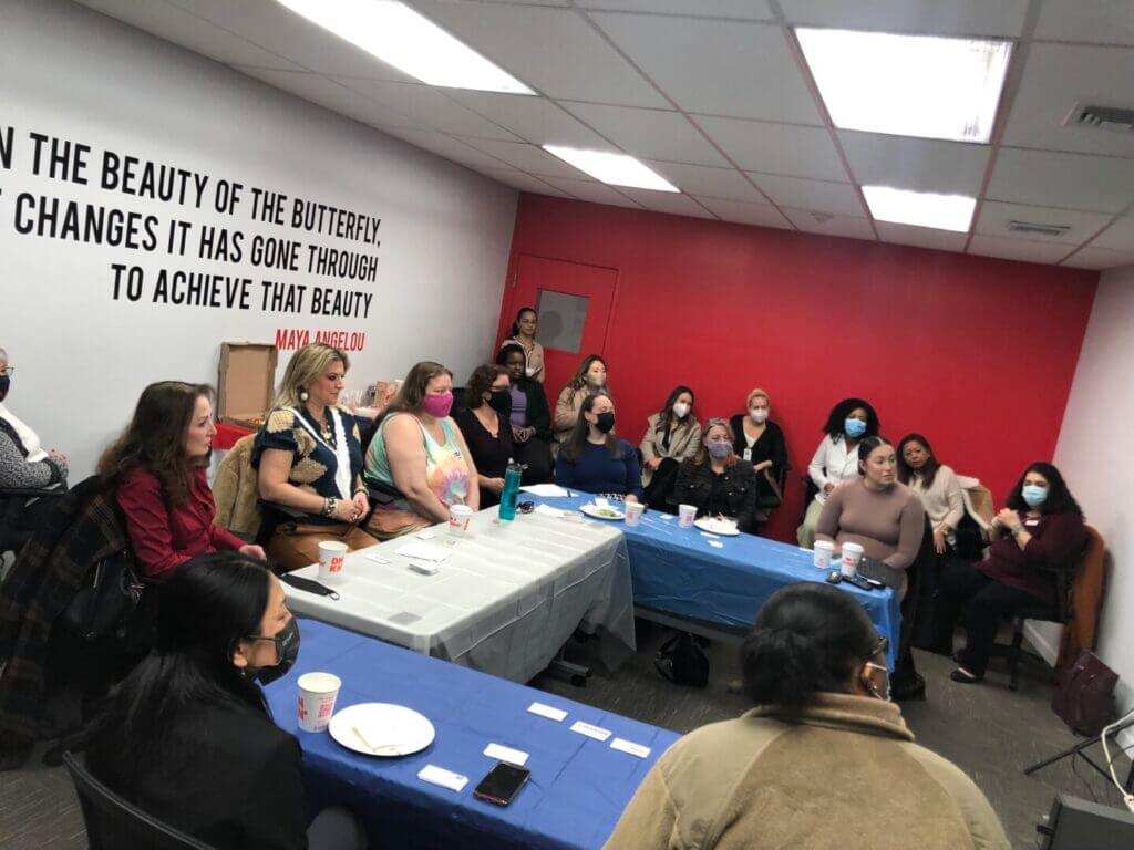 Women in a meeting room discussing, seated around tables with a Maya Angelou quote on the wall. Diverse group, masks, engaging conversation.