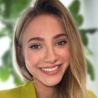 Smiling woman with blonde hair in yellow blazer, green leaves in background.