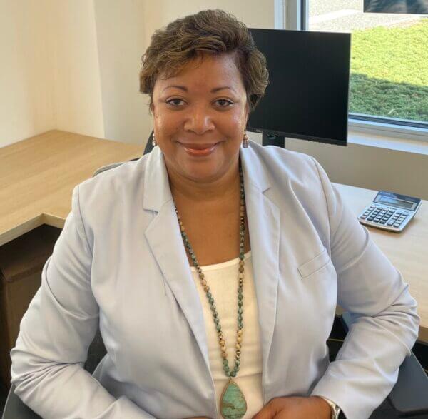 Andresa Moody smiling in office, wearing a light blue blazer and turquoise necklace, with a computer and calculator on the desk.