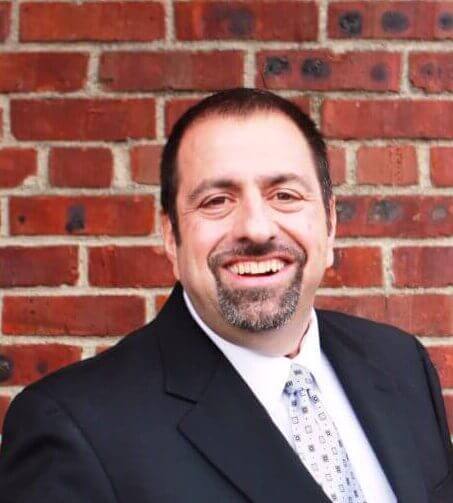 Man in a suit smiling in front of a brick wall background.