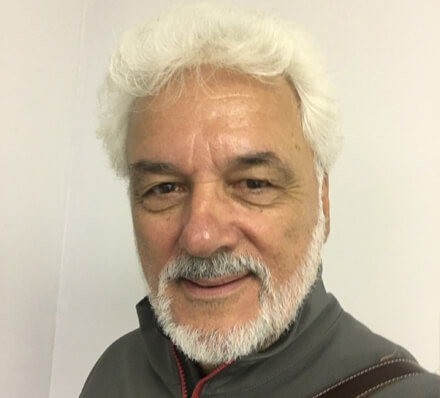 Elderly man with white hair and beard smiling, wearing a gray jacket against a plain background.